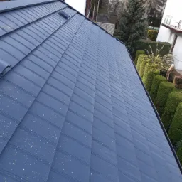 Nowy, niebieski dach z blachy trapezowej z widocznym kominkiem wentylacyjnym na tle zadbanego ogrodu i zabudowy jednorodzinnej.