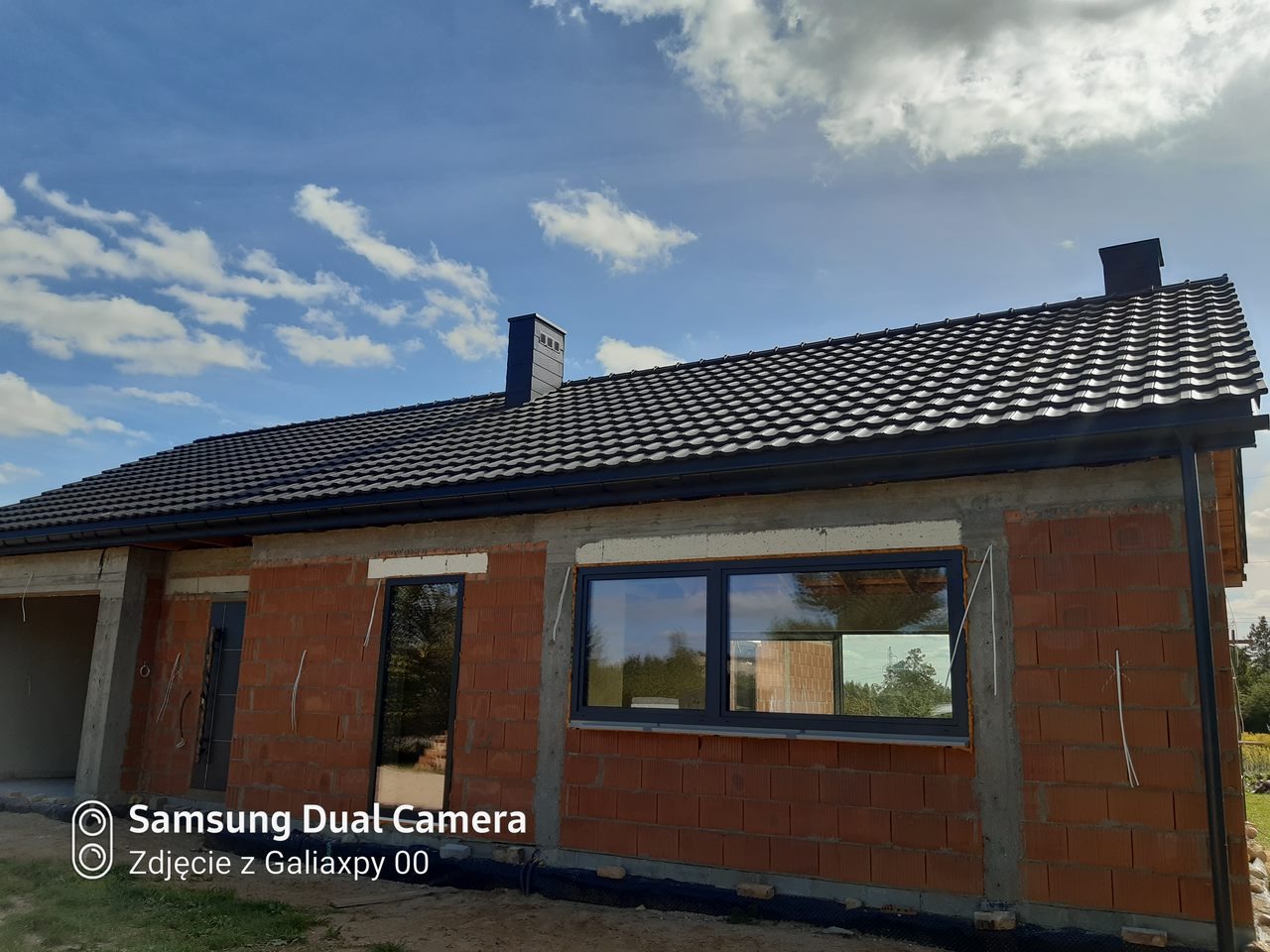 Dom w trakcie budowy z czarnym dachem z dachówki ceramicznej, widoczne okna z szarymi ramami, ściany z czerwonej cegły i komin z cegły, błękitne niebo z chmurami w tle.