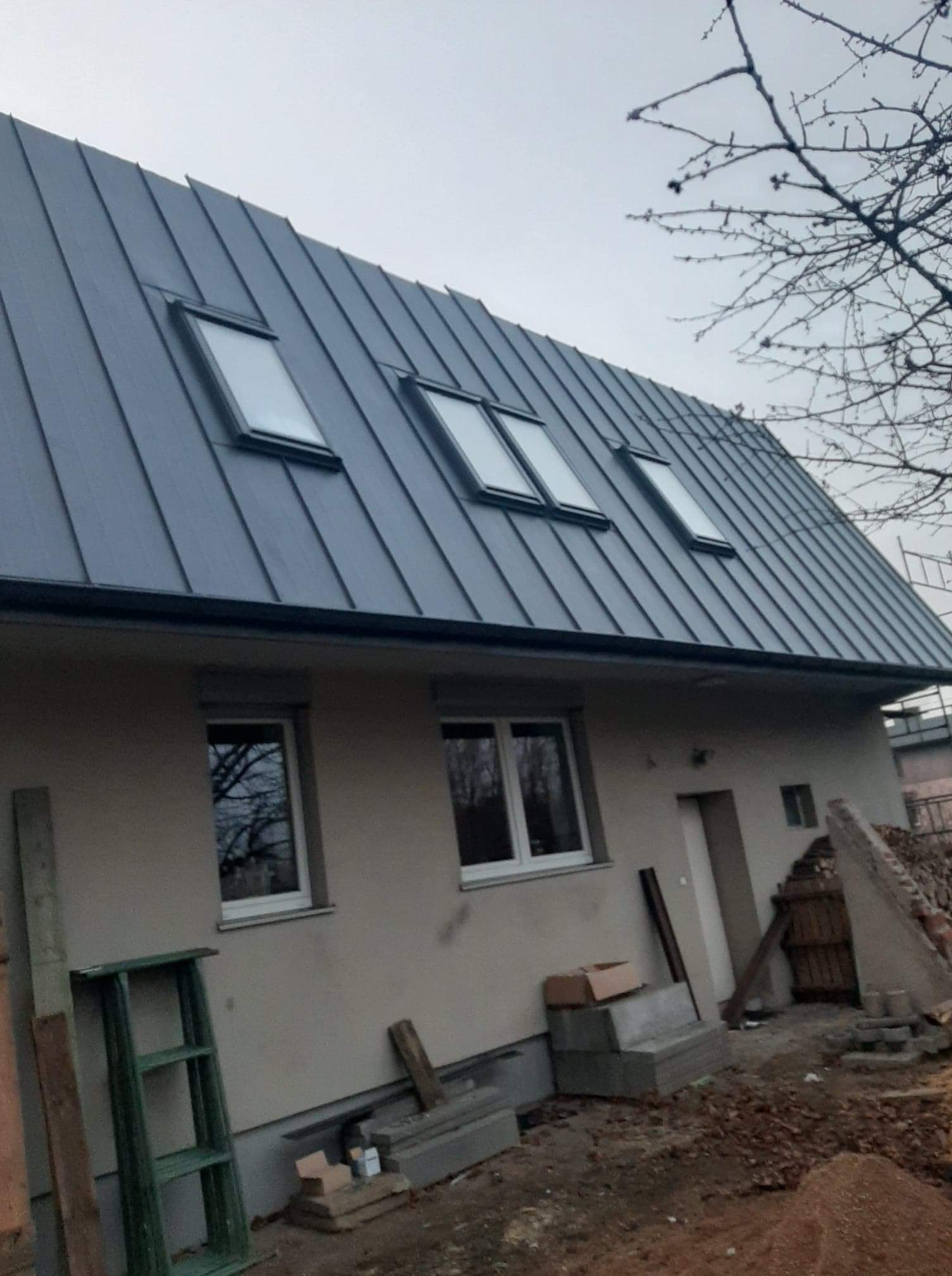 Dom z nowym, szarym dachem z blachy na rąbek stojący i trzema oknami dachowymi; widoczne rusztowania i materiały budowlane.