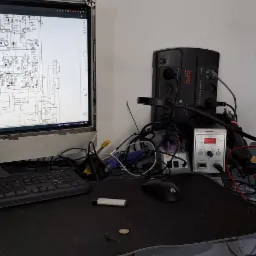 Stanowisko pracy elektronika: schemat na monitorze iMac, multimetr UNI-T wskazujący 0.00, stacja lutownicza, zasilacz awaryjny APC i klawiatura Steelseries na czarnym biurku.