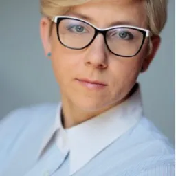 Portret kobiety w okularach z białą oprawką, w błękitnej koszuli z białym kołnierzykiem, patrzącej wprost.