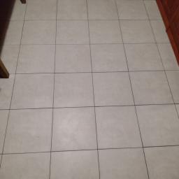 Wojciech Łada - Beżowa podłoga wyłożona kwadratowymi płytkami ceramicznymi z ciemnymi fugami, fragmenty mebli w tle.