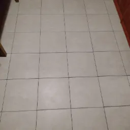 Beżowa podłoga wyłożona kwadratowymi płytkami ceramicznymi z ciemnymi fugami, fragmenty mebli w tle.