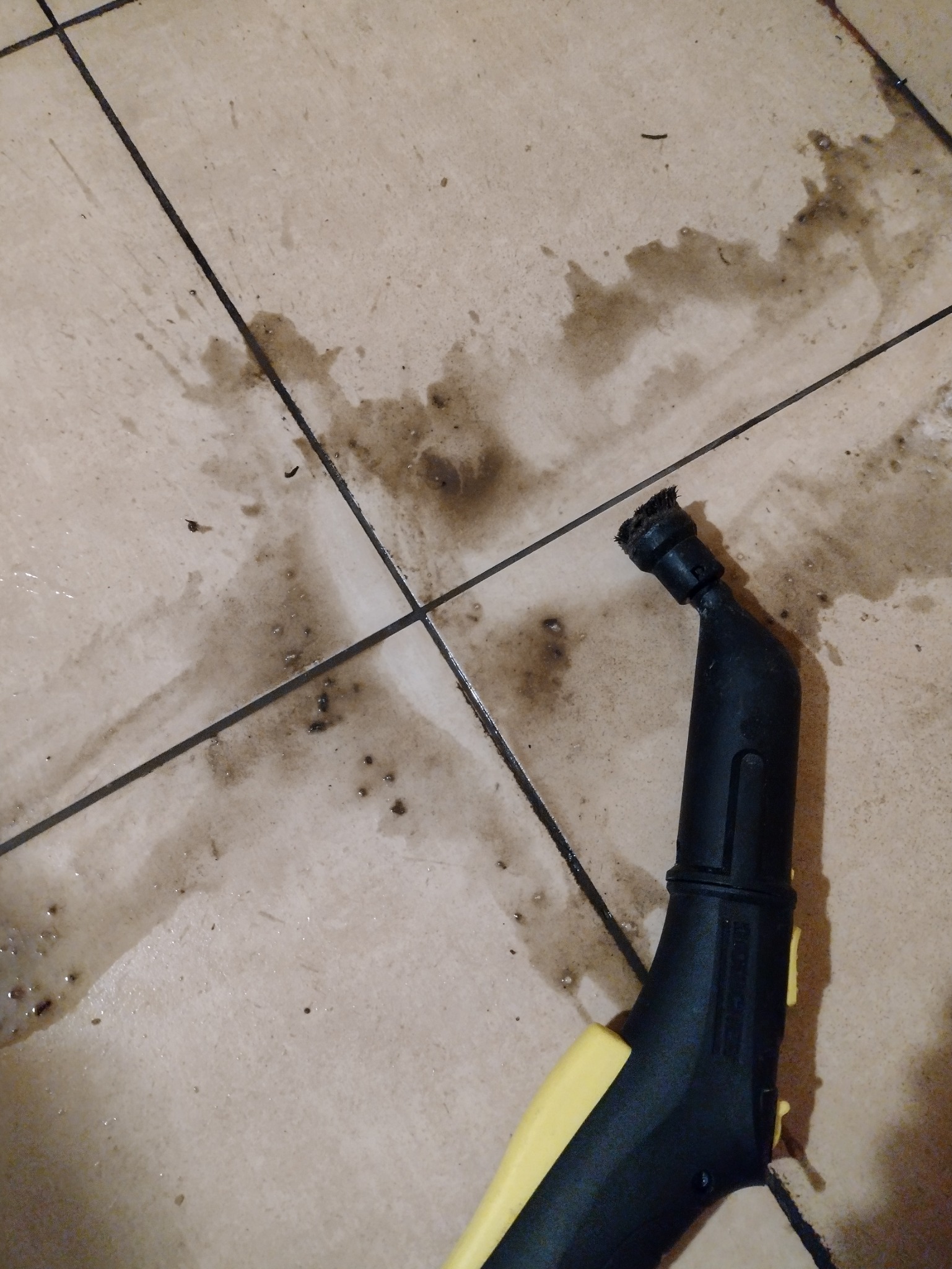 Czyszczenie fug i płytek podłogowych parownicą z czarną dyszą i żółtym uchwytem, widoczne zabrudzenia i woda na jasnych płytkach.