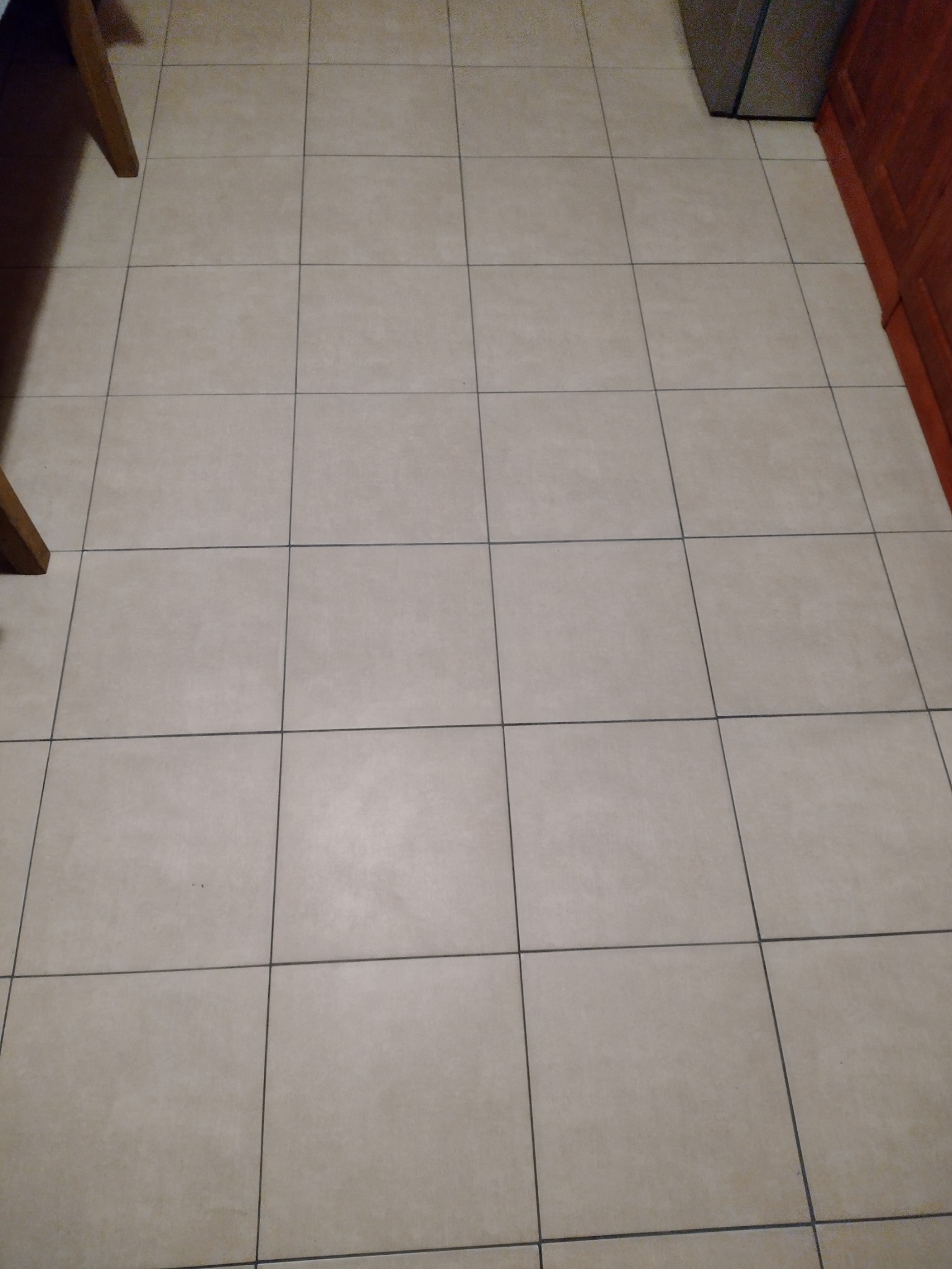 Beżowa podłoga wyłożona kwadratowymi płytkami ceramicznymi z ciemnymi fugami, fragmenty mebli w tle.