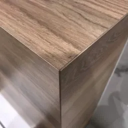 Detal narożnika szafki łazienkowej z drewnopodobnym fornirem, widoczna precyzja wykonania i dopasowanie krawędzi, na tle marmurowej ściany.