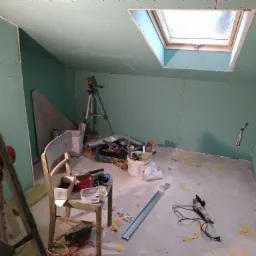 Poddasze w trakcie wykańczania płytami karton-gips z widocznym oknem dachowym, laserowym niwelatorem na statywie, narzędziami i przewodami na podłodze oraz starym krzesłem.