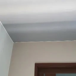 Fragment wnętrza: gładko pomalowany sufit w odcieniach szarości, jasna ściana i fragment brązowej ramy drzwi, widoczne pod kątem.