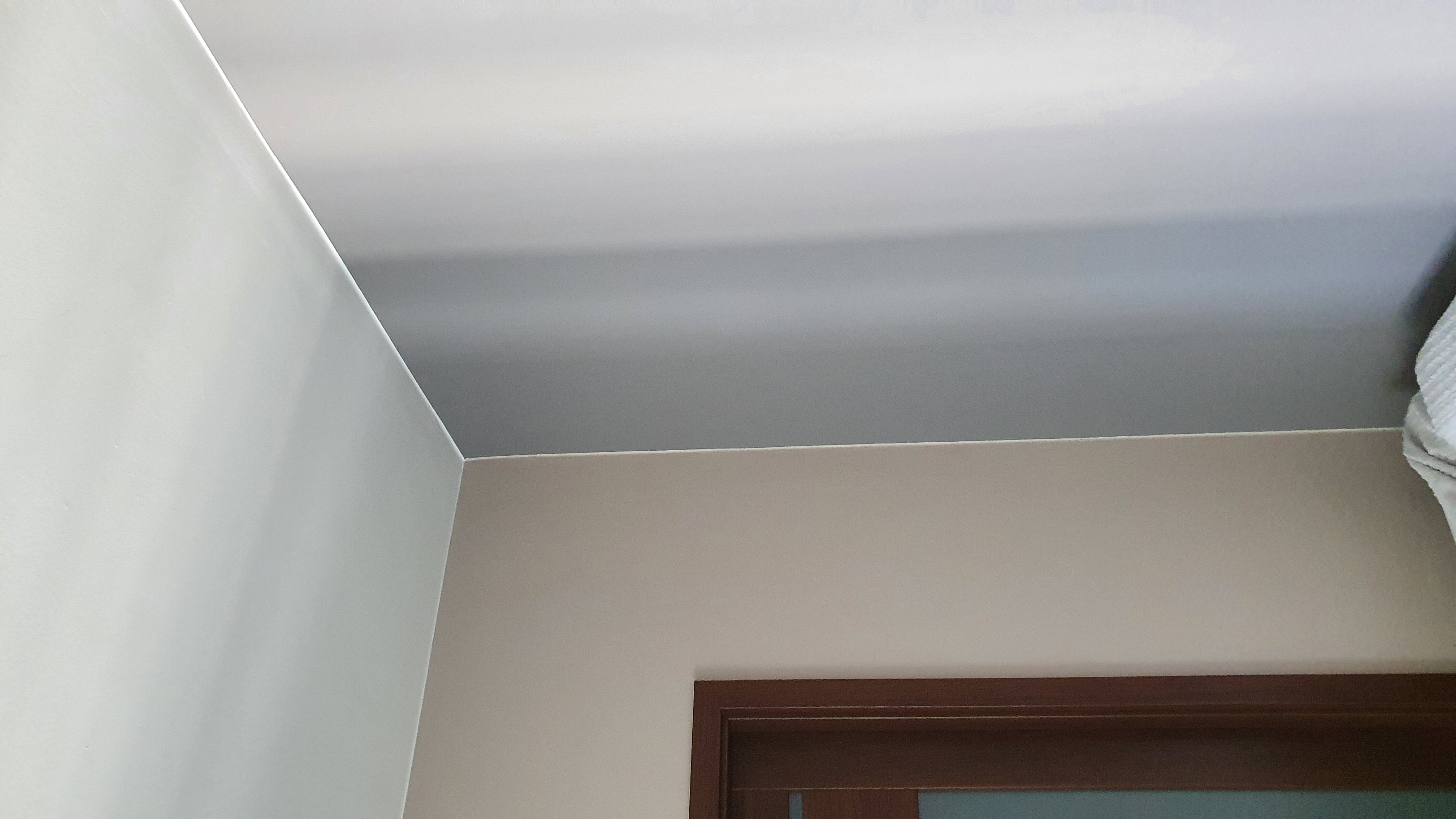 Fragment wnętrza: gładko pomalowany sufit w odcieniach szarości, jasna ściana i fragment brązowej ramy drzwi, widoczne pod kątem.