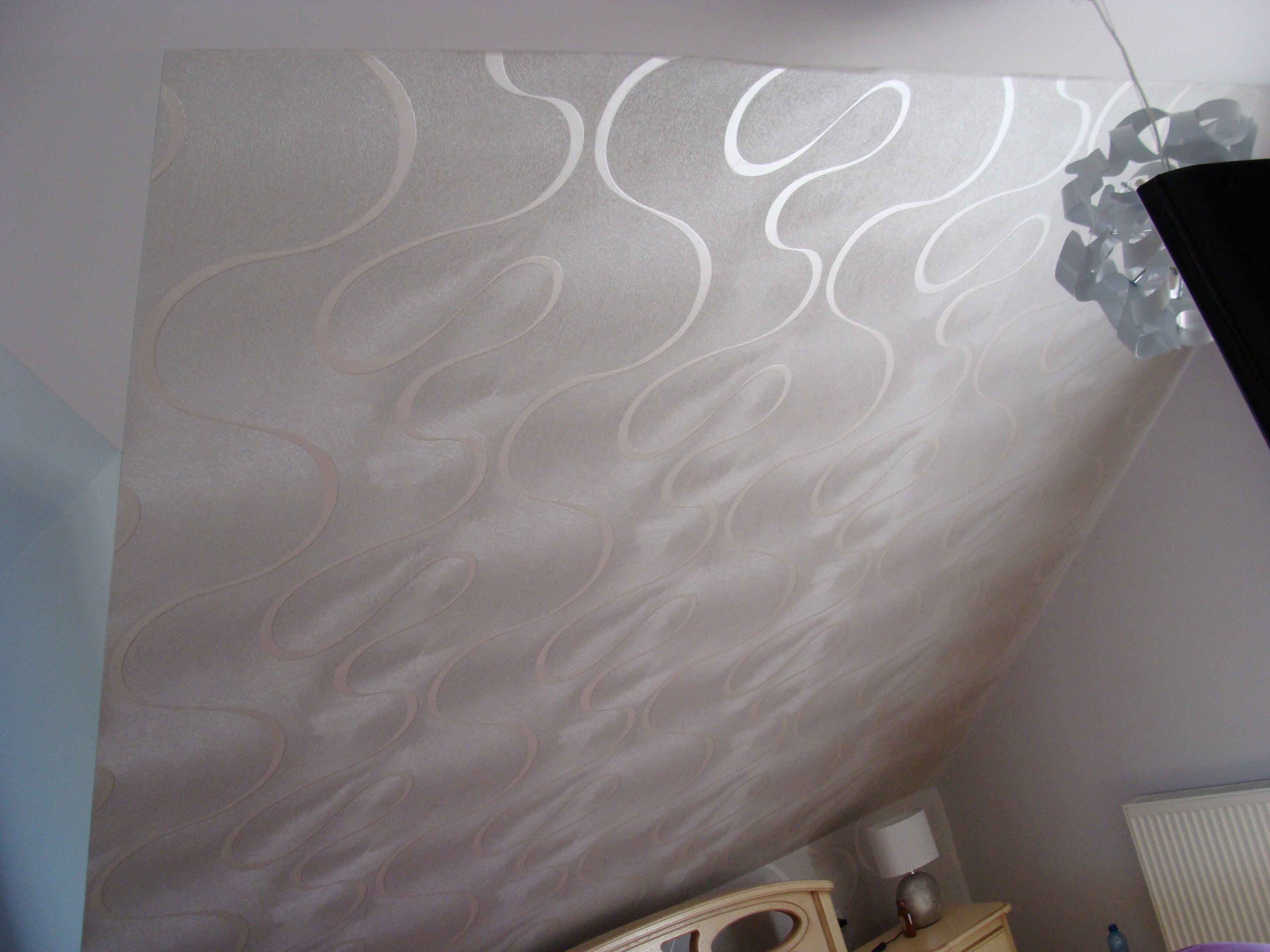 Ściana skośna w pokoju na poddaszu pokryta tapetą w odcieniach beżu z delikatnym, falistym wzorem, widoczny fragment lampy sufitowej i mebli.