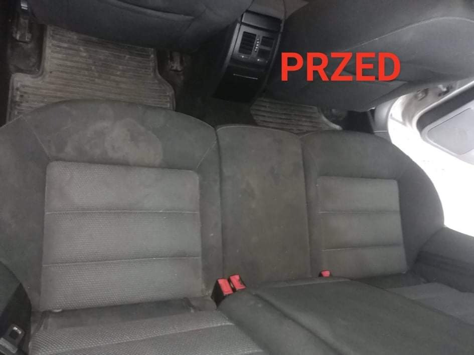 Brudna, szara tylna kanapa samochodowa z plamami i napisem 'PRZED' w prawym górnym rogu, widoczne zagłówki i fragment podłogi z dywanikiem