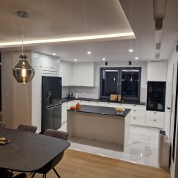 Ewelina Galwiaczek Meble Ni-Na - Nowoczesna, jasna kuchnia z wyspą i czarnymi blatami w Radziechowicach Pierwszych, z widoczną lampą wiszącą w stylu retro i elementami dekoracyjnymi na wyspie.