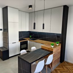 Ewelina Galwiaczek Meble Ni-Na - Nowoczesna kuchnia z białymi szafkami, blatem z imitacją drewna i czarnym panelem ściennym z marmurkowym wzorem, uzupełniona czarnym stołem z białymi krzesłami i minimalistycznym oświetleniem.