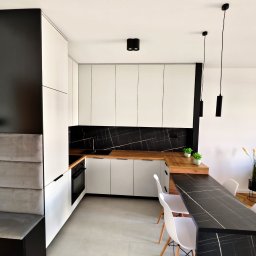 Ewelina Galwiaczek Meble Ni-Na - Nowoczesna kuchnia z białymi szafkami, drewnianym blatem, czarnym panelem ściennym imitującym marmur i czarnym barkiem z krzesłami, oświetlona wiszącymi lampami tubami.