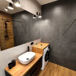 Nowoczesna łazienka z drewnianym blatem pod umywalką nablatową, szarą płytką na ścianie i drewnianą podłogą. W tle widoczny prysznic z czarną armaturą odbijający się w lustrze.