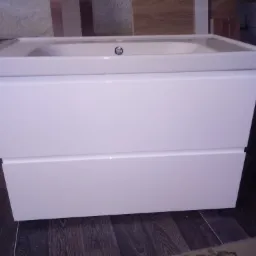 Biała szafka pod umywalkę z dwiema szufladami, widoczna umywalka z chromowanym korkiem, tło stanowią materiały budowlane i surowa ściana.