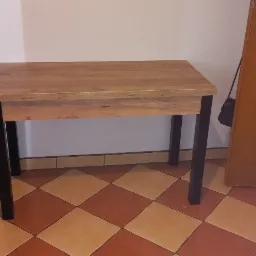 Prostokątny stolik z blatem imitującym jasne drewno i czarnymi nogami, ustawiony na podłodze w brązowo-pomarańczową szachownicę, obok ściany i drewnianych drzwi z owalnym przeszkleniem.