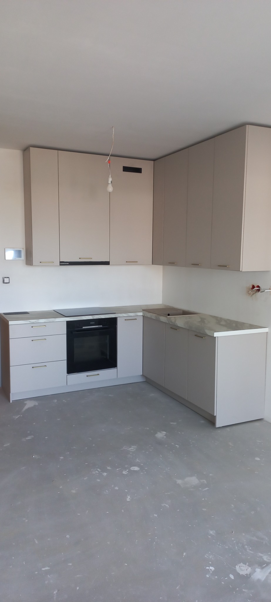 Jasna kuchnia w trakcie montażu, widoczne szafki górne i dolne z szarymi blatami, płyta indukcyjna i piekarnik, surowy stan podłogi, brakujące oświetlenie.