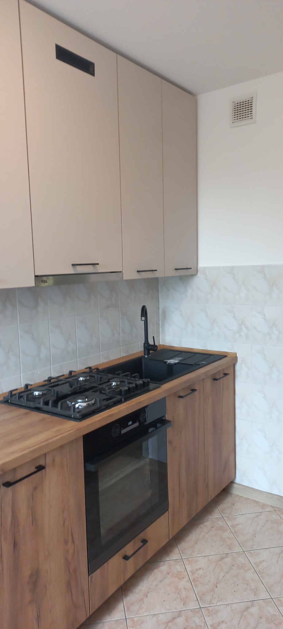 Fragment zabudowy kuchennej z beżowymi szafkami wiszącymi, blatem imitującym drewno, czarnym zlewem i płytą gazową, z piekarnikiem w zabudowie pod płytą, na tle marmurowych płytek ściennych.