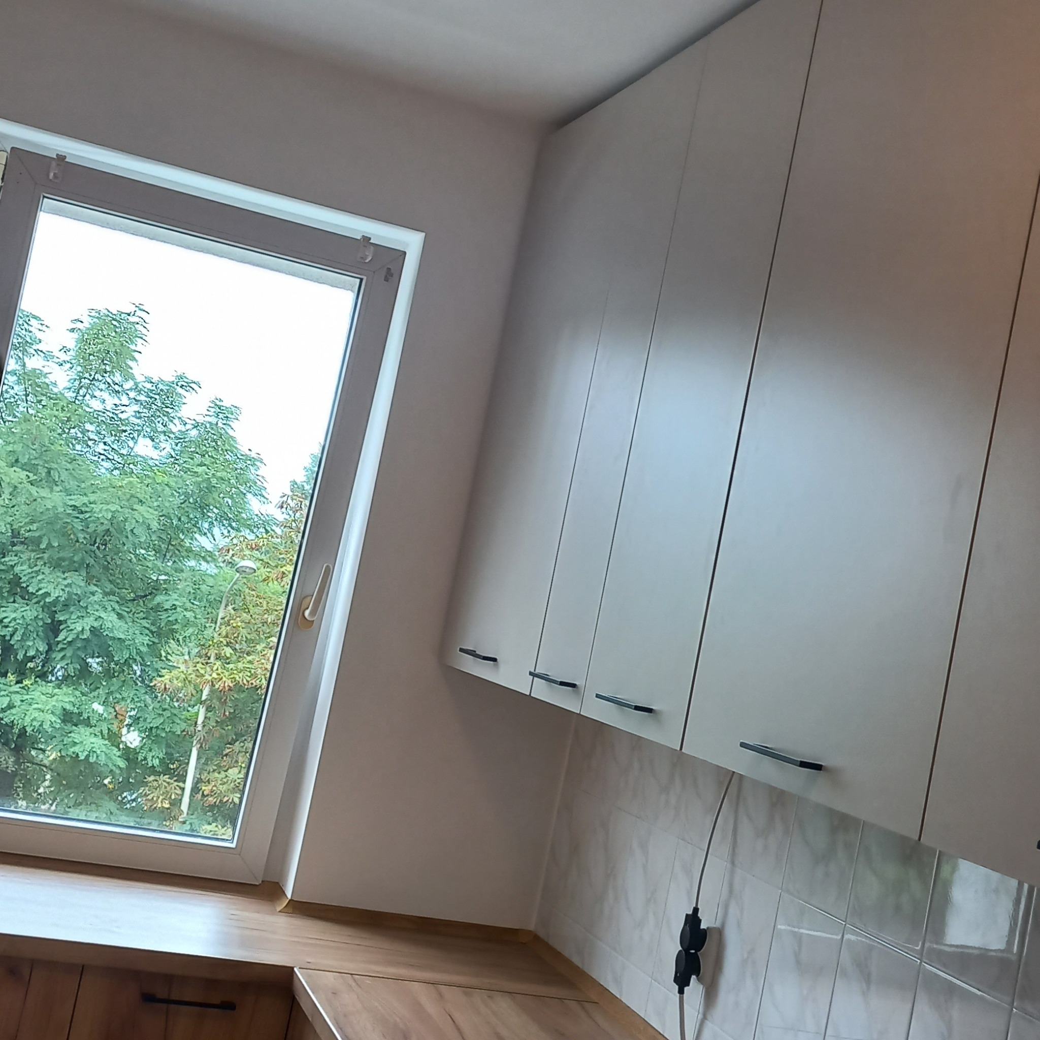 Nowoczesna kuchnia z białymi szafkami wiszącymi z czarnymi uchwytami, drewnianym blatem i widokiem na zieleń za oknem, z widocznym gniazdkiem elektrycznym na ścianie z marmurkowymi płytkami.