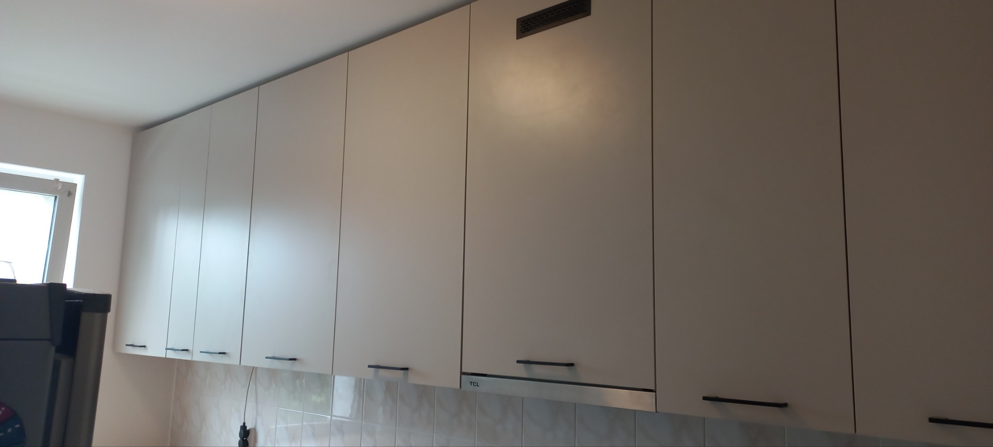 Rząd górnych szafek kuchennych w odcieniach bieli z czarnymi, minimalistycznymi uchwytami, zamontowanych nad płytkami ściennymi, z widocznym fragmentem okna i lodówki po lewej stronie.