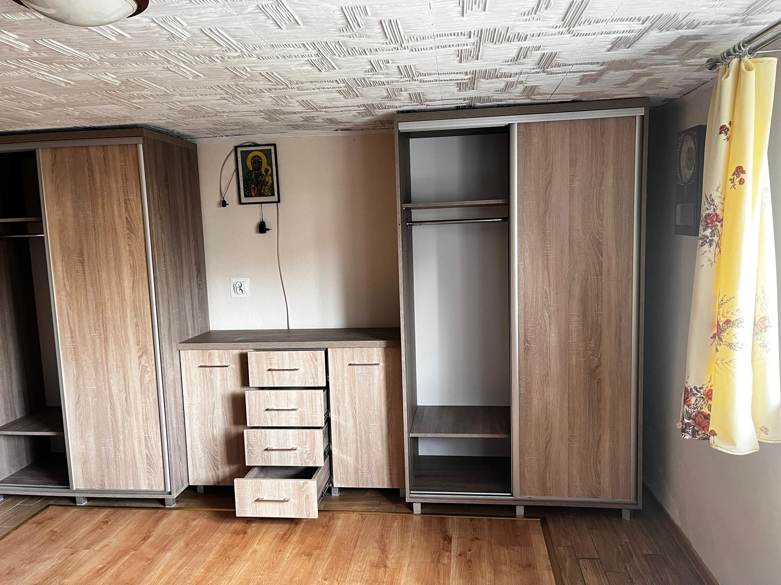 Zestaw mebli: szafa przesuwna z półkami i drążkiem, komoda z szufladami (jedna otwarta), całość w odcieniach brązu, na ścianie ikona.