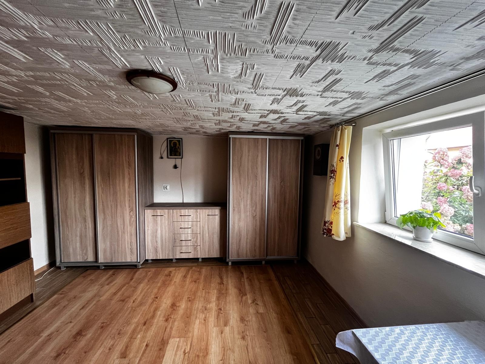 Pokój z dwiema szafami przesuwnymi z drewnopodobnymi frontami, komodą z szufladami, wzorzystym sufitem i oknem z kwiatami za szybą.