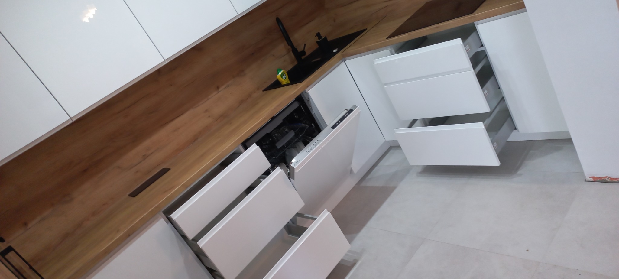 Nowoczesna kuchnia z białymi frontami i drewnianym blatem, widoczne otwarte szuflady i zmywarka, zlew z czarną armaturą i płynem do naczyń.