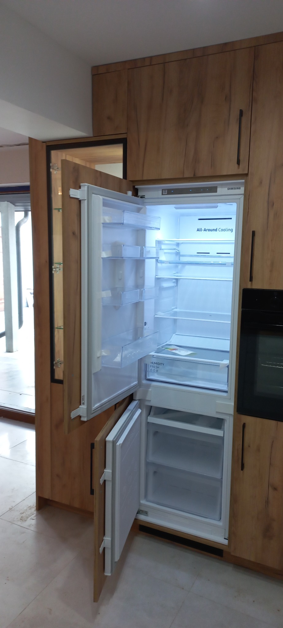 Wbudowana lodówka Samsung z otwartymi drzwiami, umieszczona w drewnianej zabudowie kuchennej z ciemnymi uchwytami, obok zabudowany piekarnik, widoczne szklane półki w szafce po lewej stronie.