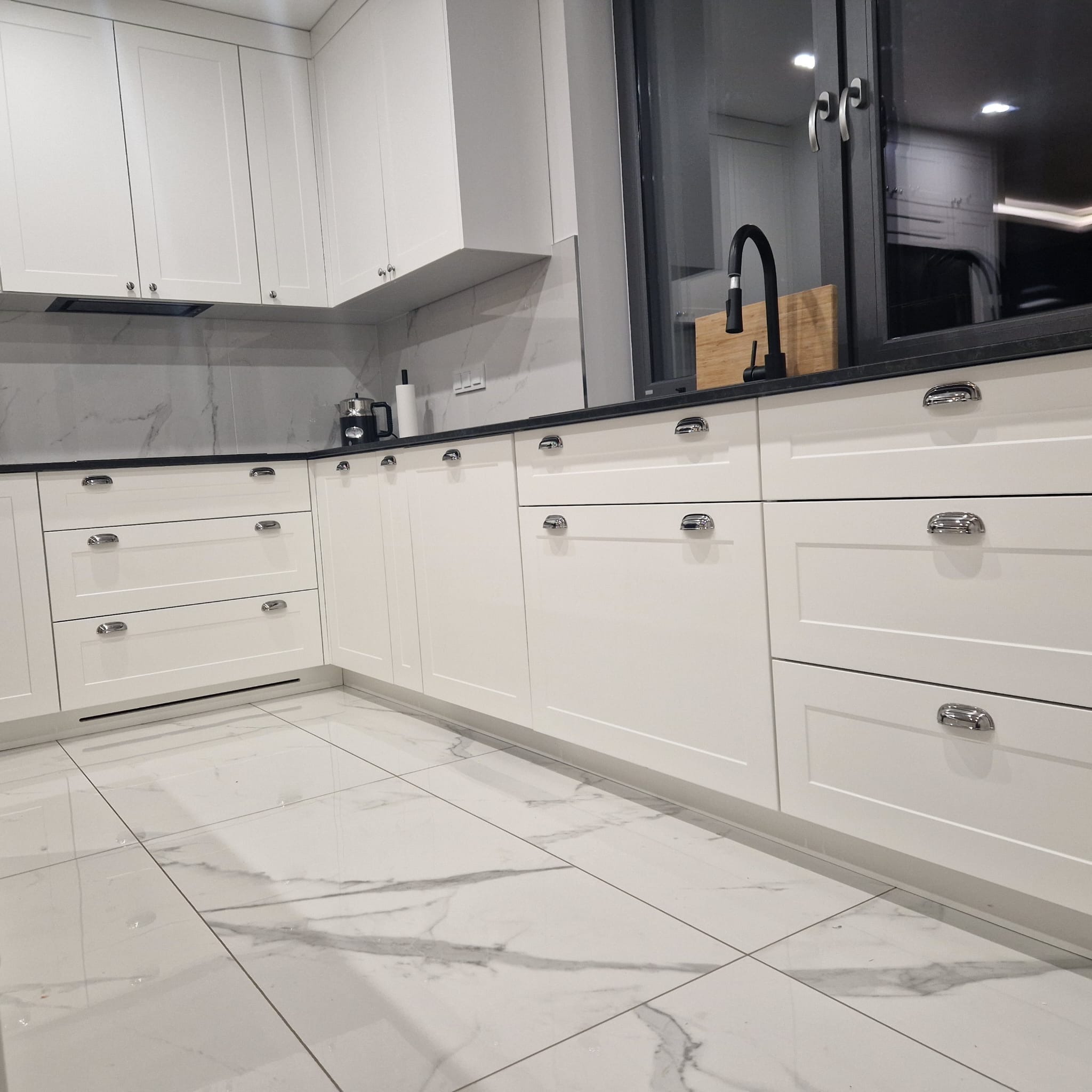 Nowoczesna, biała kuchnia z marmurowym blatem i podłogą, chromowane uchwyty, czarna bateria kuchenna i okno z czarną ramą.