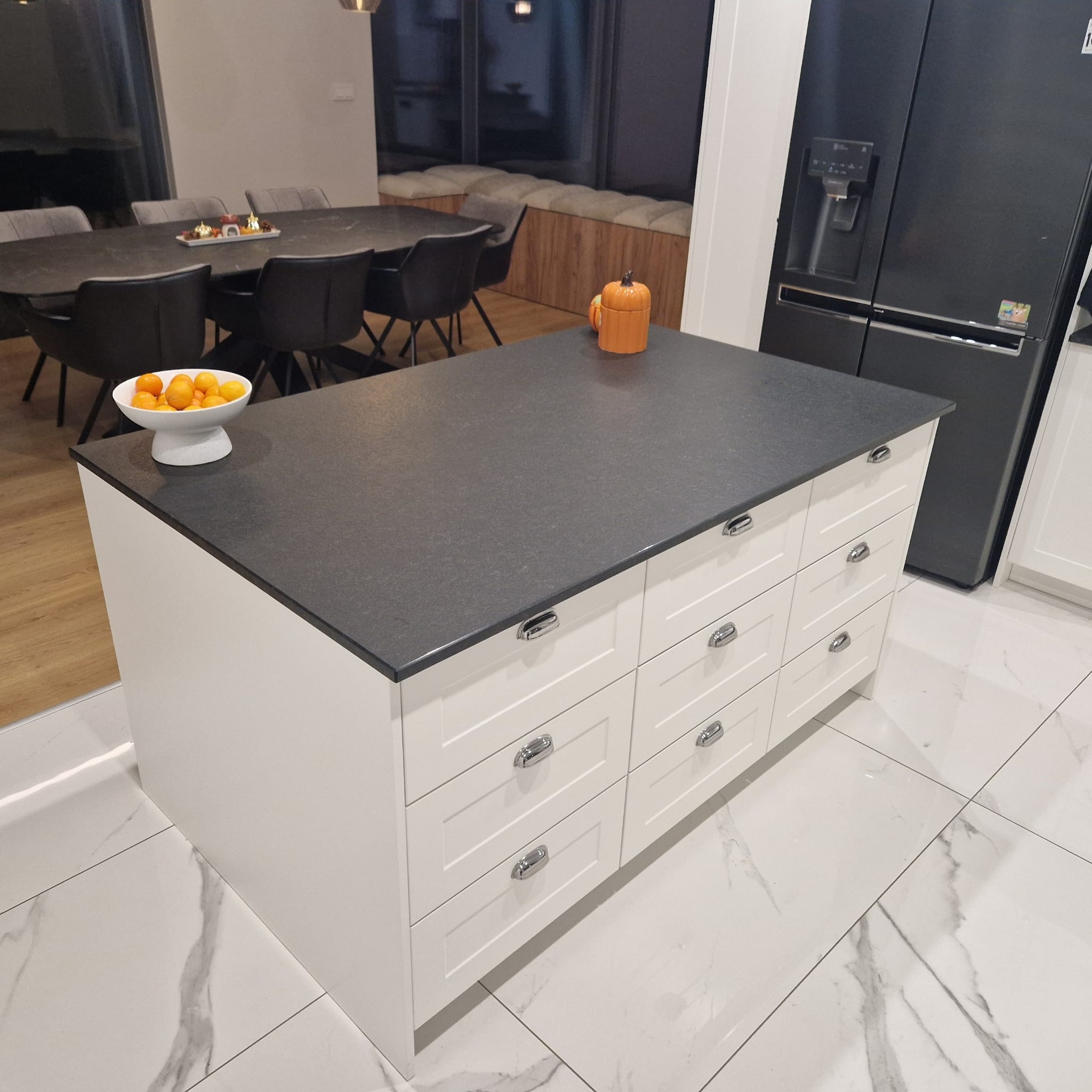 Wyspa kuchenna z białymi frontami szafek z szufladami i ciemnoszarym blatem w nowoczesnej kuchni, na blacie miska z pomarańczami i ceramiczna dynia.