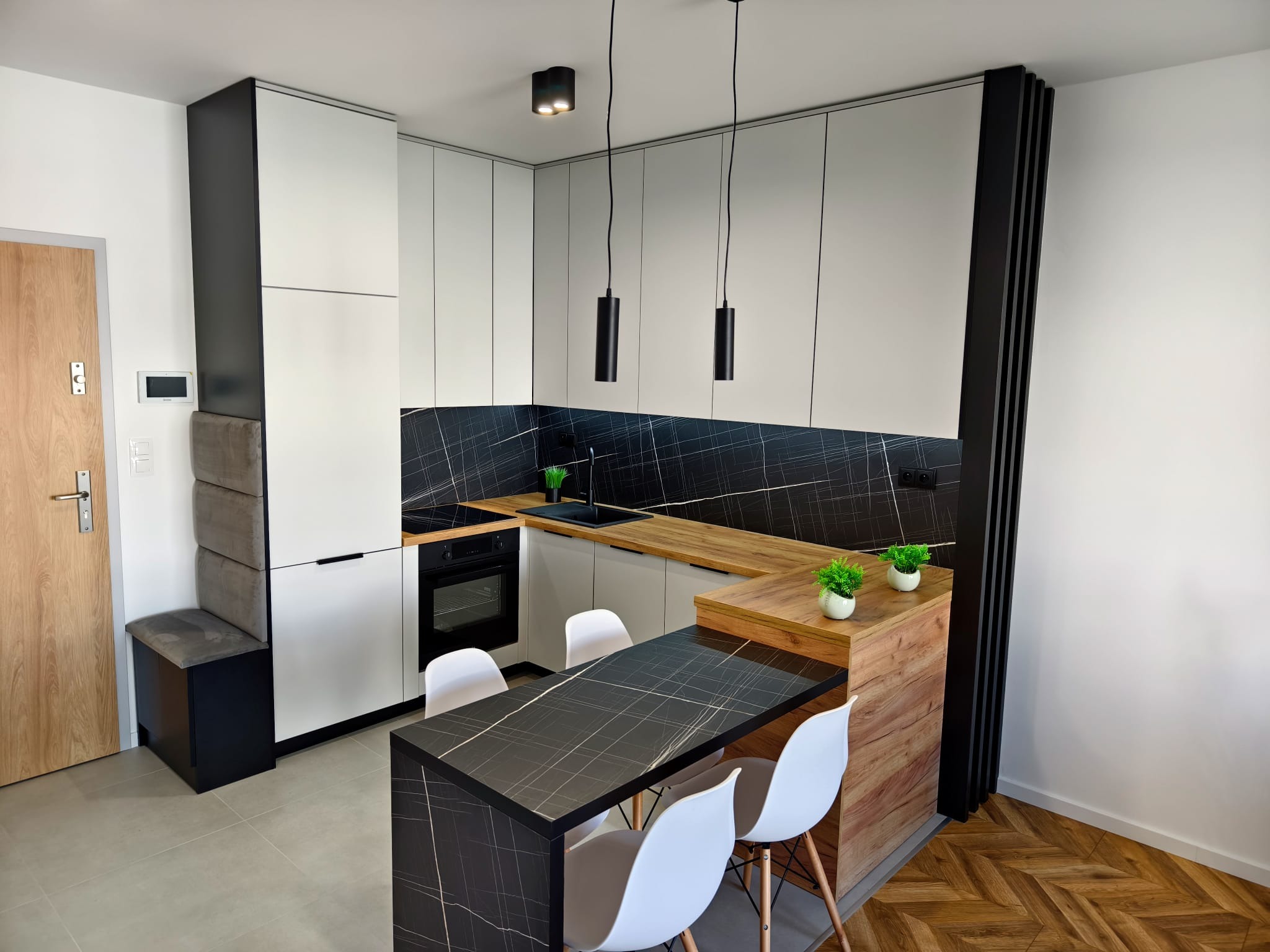 Nowoczesna kuchnia z białymi szafkami, blatem z imitacją drewna i czarnym panelem ściennym z marmurkowym wzorem, uzupełniona czarnym stołem z białymi krzesłami i minimalistycznym oświetleniem.