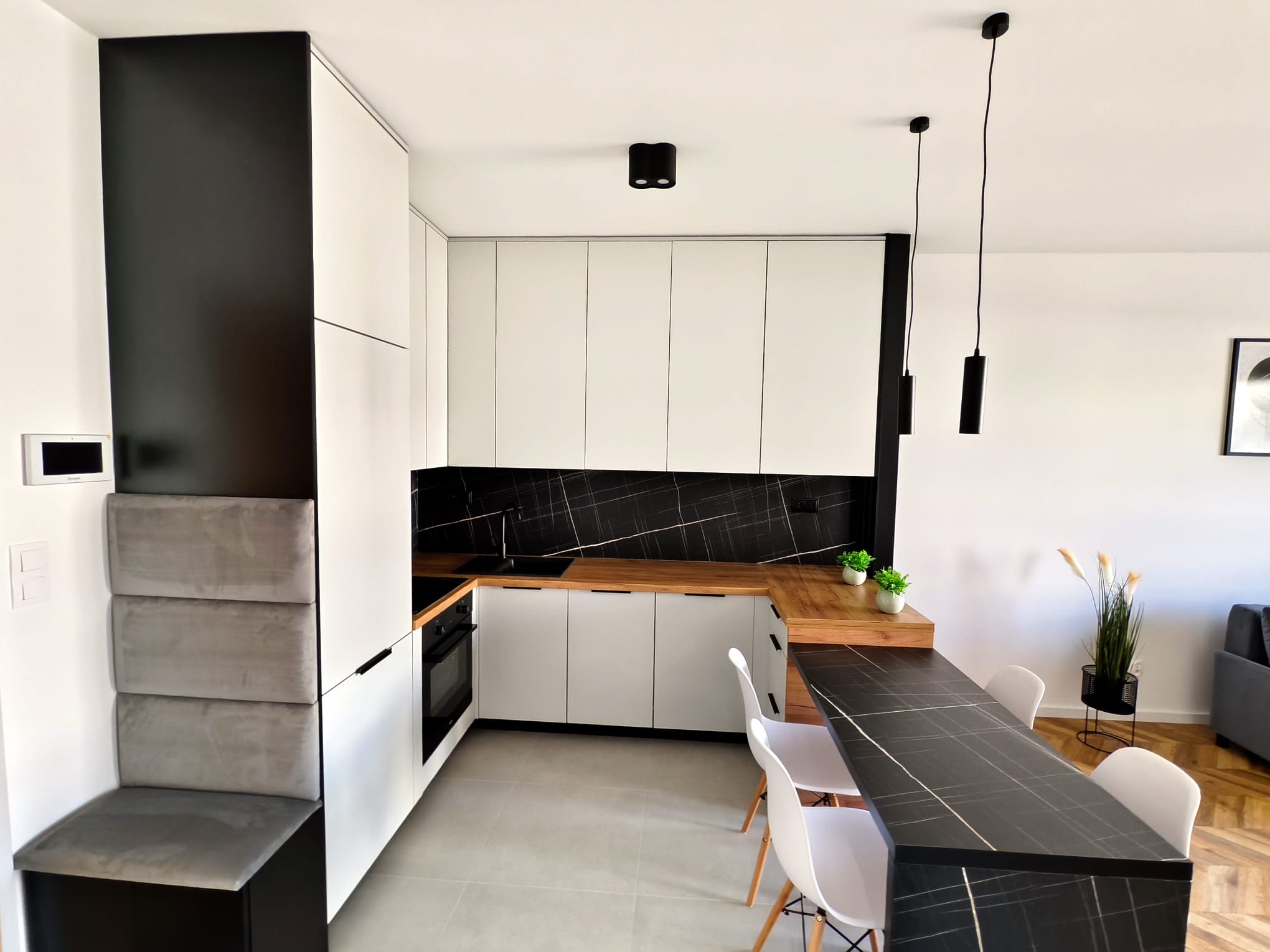 Nowoczesna kuchnia z białymi szafkami, drewnianym blatem, czarnym panelem ściennym imitującym marmur i czarnym barkiem z krzesłami, oświetlona wiszącymi lampami tubami.