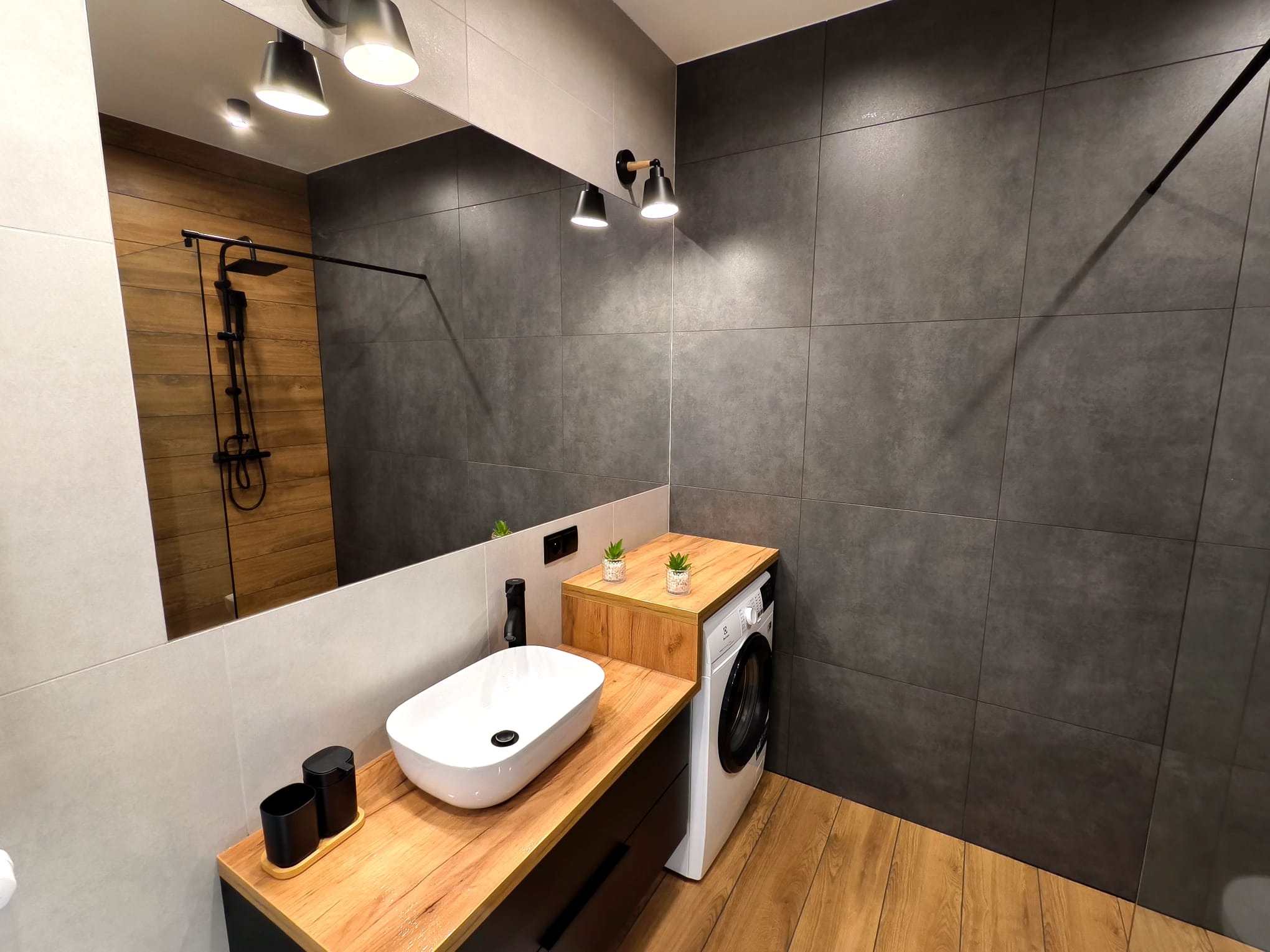 Nowoczesna łazienka z drewnianym blatem pod umywalką nablatową, szarą płytką na ścianie i drewnianą podłogą. W tle widoczny prysznic z czarną armaturą odbijający się w lustrze.