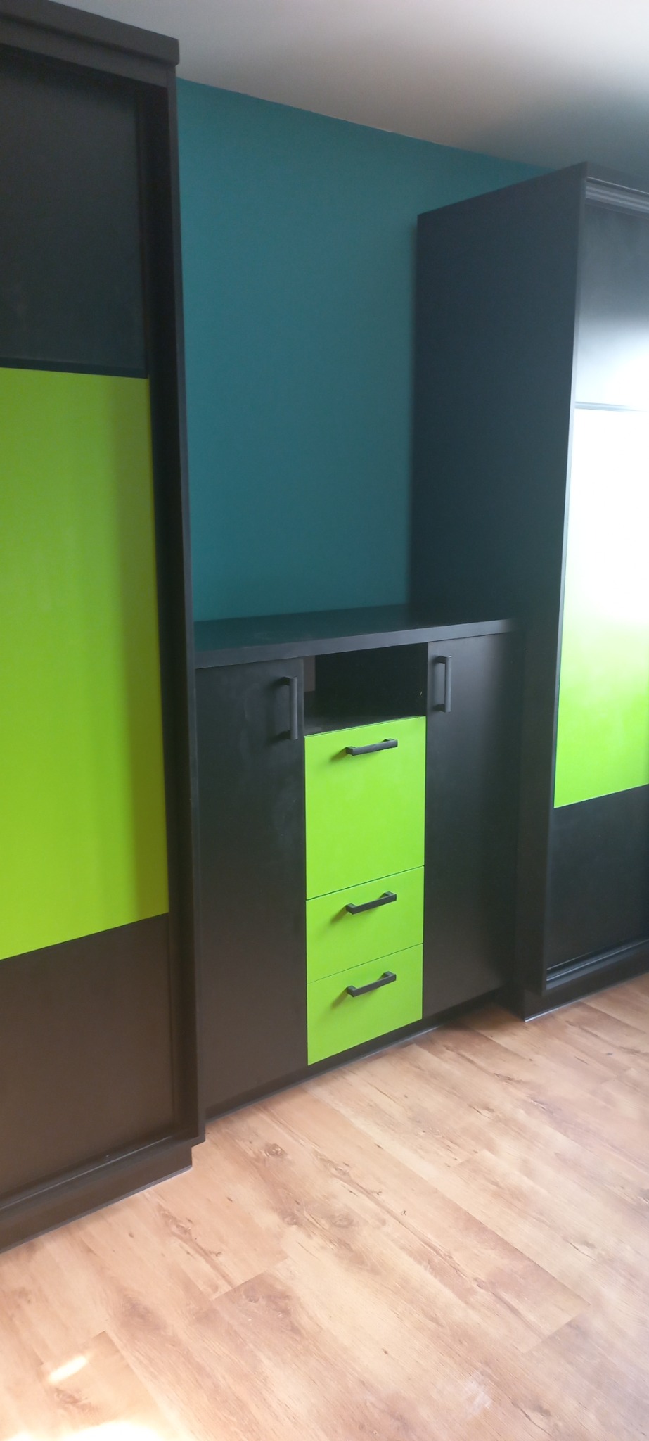 Ciemnoszara szafa z przesuwnymi drzwiami i szafką z trzema jaskrawozielonymi szufladami, na drewnianej podłodze, na tle turkusowej ściany.