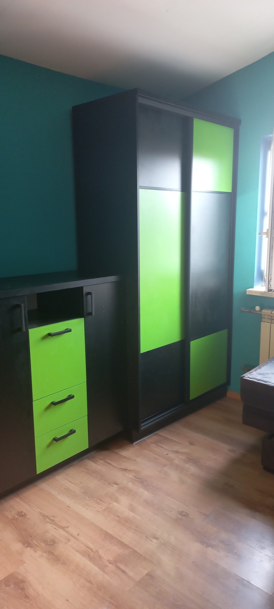 Ciemna szafa przesuwna z zielonymi akcentami i komoda z szufladami w pokoju z turkusowymi ścianami i drewnianą podłogą.