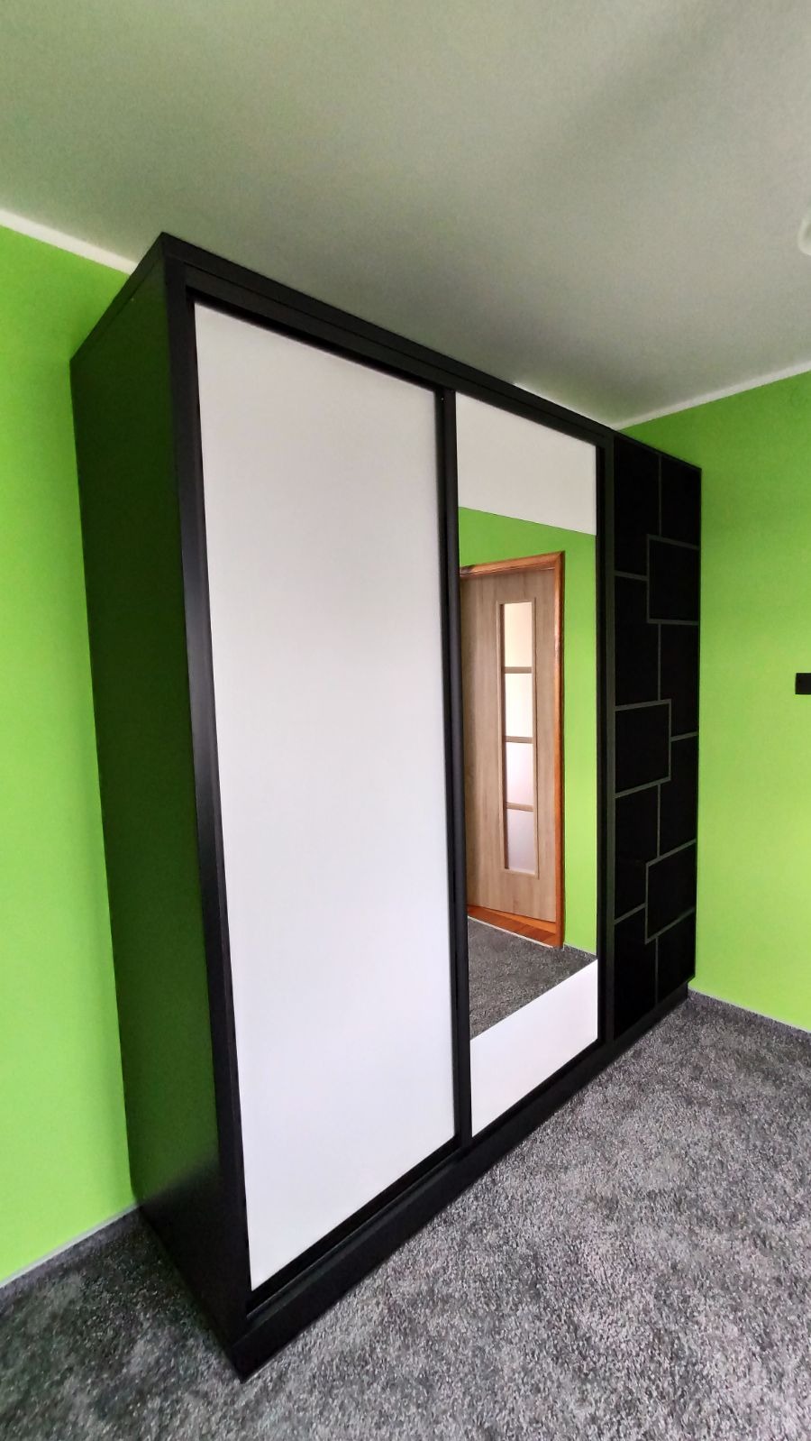Nowoczesna szafa przesuwna z białymi frontami i lustrem, czarna rama, dekoracyjny panel boczny z czarnych kwadratów, widok na drzwi w odbiciu lustra, zielona ściana w tle.