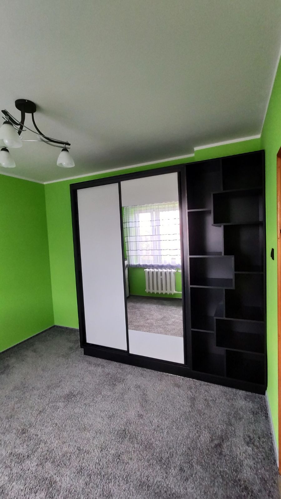 Czarna szafa przesuwna z białymi frontami i lustrem w pokoju z zielonymi ścianami i szarym dywanem, widoczna także część regału z półkami po prawej stronie szafy.