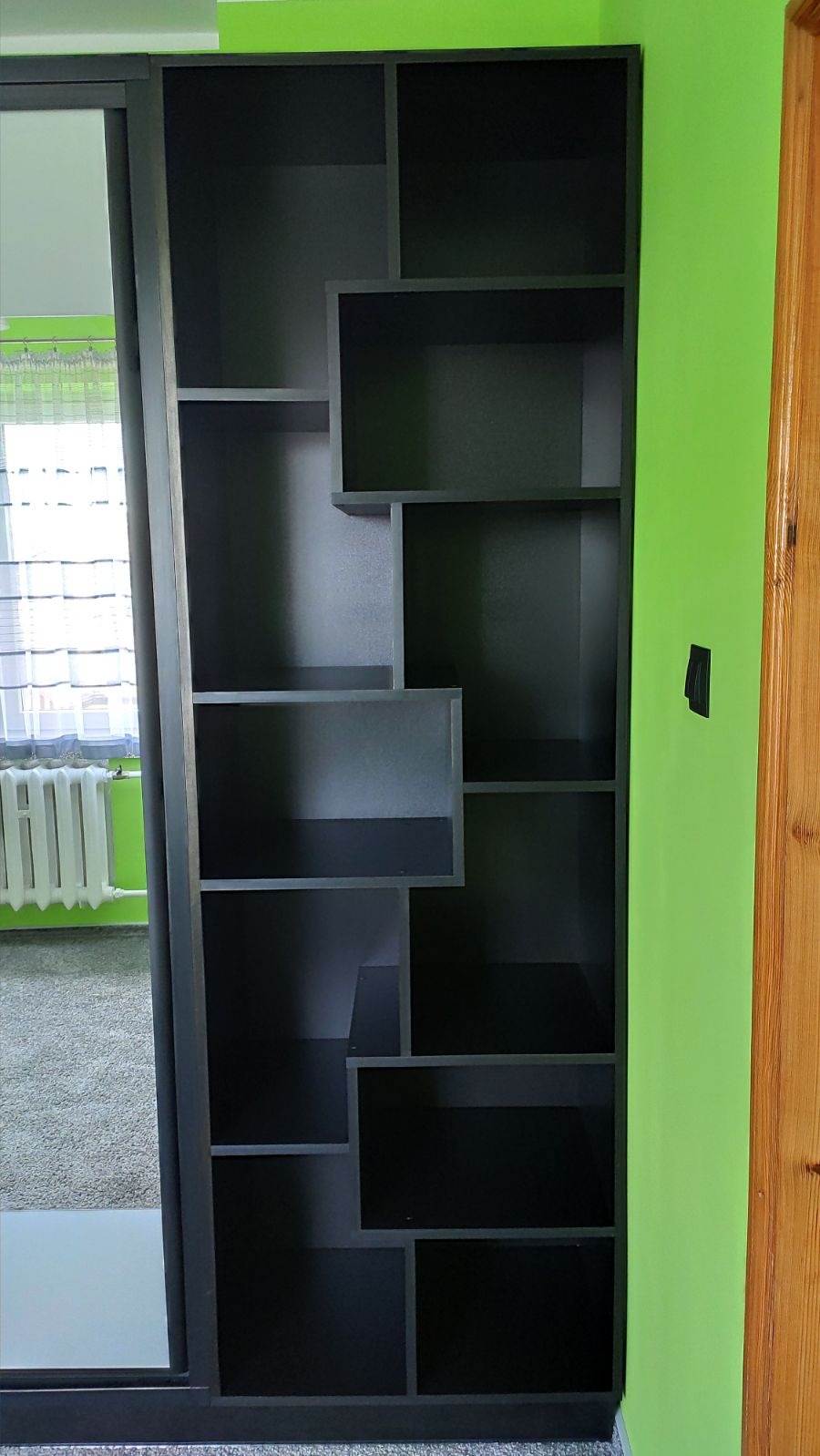 Ciemnoszara szafa wnękowa z przesuwnymi drzwiami lustrzanymi i asymetrycznymi półkami, na tle limonkowo-zielonej ściany i drewnianych drzwi.