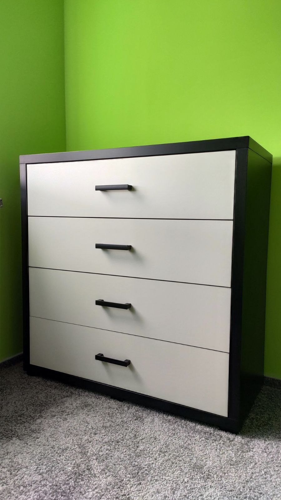 Czteroszuflowa komoda z białymi frontami i czarną ramą, ustawiona na szarym dywanie na tle jaskrawozielonej ściany.