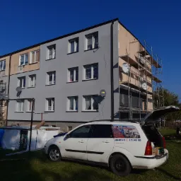 Blok mieszkalny w trakcie prac elewacyjnych, część budynku pomalowana na szaro, druga w kolorze beżowym, widoczne rusztowania, obok zaparkowany samochód z reklamą usług budowlanych.