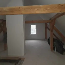 Poddasze w trakcie remontu, widoczne drewniane elementy konstrukcyjne dachu, fragment ściany z włącznikami i okno w tle, na podłodze resztki materiałów budowlanych.