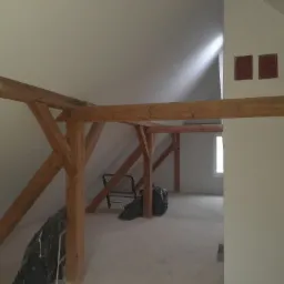 Surowe drewniane belki stropowe w jasnym, surowym wnętrzu poddasza, widoczne worki ze śmieciami i krzesło, trzy kwadratowe wgłębienia w kominie.