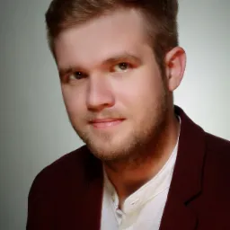 Portret młodego mężczyzny z krótką brodą, ubranego w bordową marynarkę i białą koszulę ze stójką, na jasnoszarym tle.