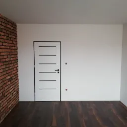 Nowoczesne wnętrze z białą ścianą, drzwiami z czarnymi akcentami, ścianą z czerwonej cegły i ciemną podłogą z paneli.