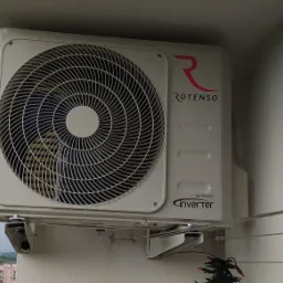 Biała zewnętrzna jednostka klimatyzacji Rotenso Inverter zamontowana na balkonie, widok na miasto w tle.