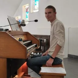 Młody mężczyzna siedzi przed organami w kościele, spoglądając w stronę kamery, widoczne witraże w tle.