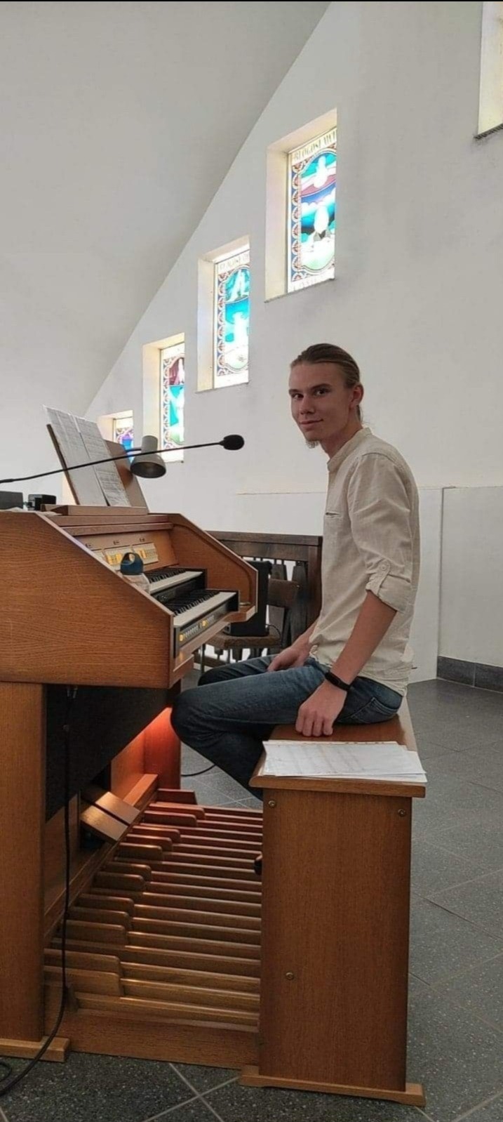 Młody mężczyzna siedzi przed organami w kościele, spoglądając w stronę kamery, widoczne witraże w tle.