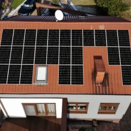 Widok z góry na dom z panelami słonecznymi na czerwonym dachu, z anteną satelitarną i kominem. Część dachu pokryta panelami, część wolna. Widoczne okno dachowe i otoczenie domu z elementami zieleni...