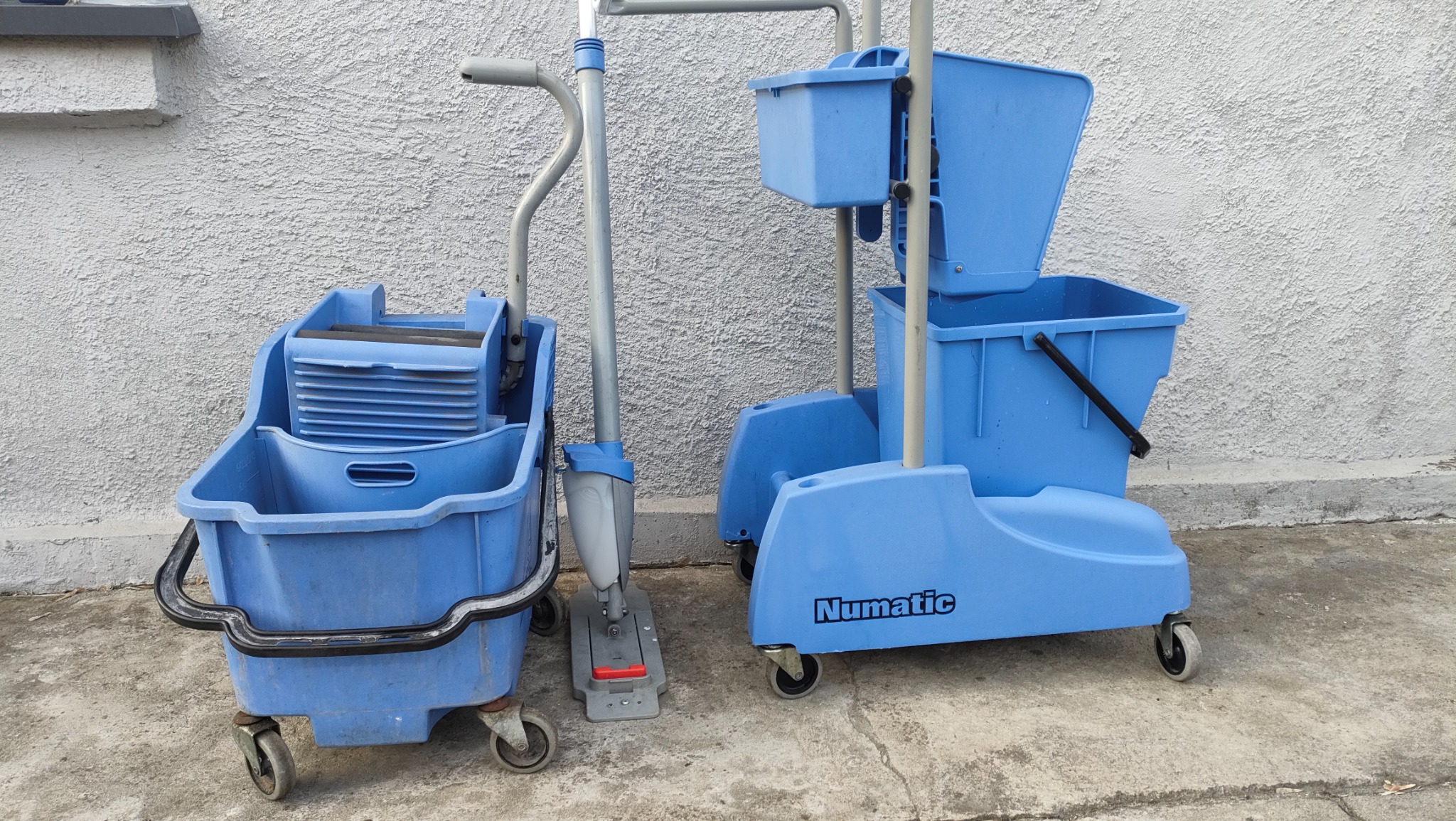 Dwa wózki do sprzątania Numatic w odcieniach niebieskiego, jeden z wyciskarką do mopa, drugi dwukomorowy, stojące na betonowej powierzchni przy białej ścianie o chropowatej fakturze.